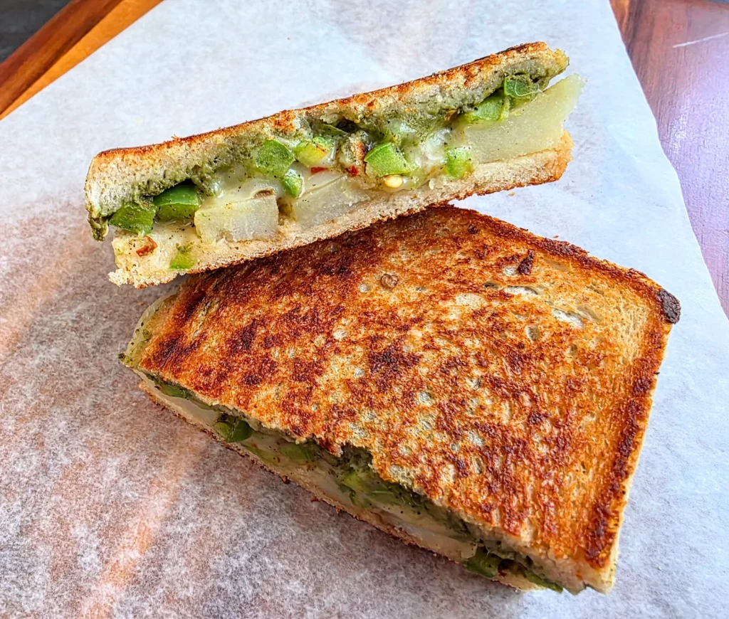 Grilled potato chutney sandwich cut in half, showing melted cheese, spiced potato slices and green chutney between toasted sourdough.