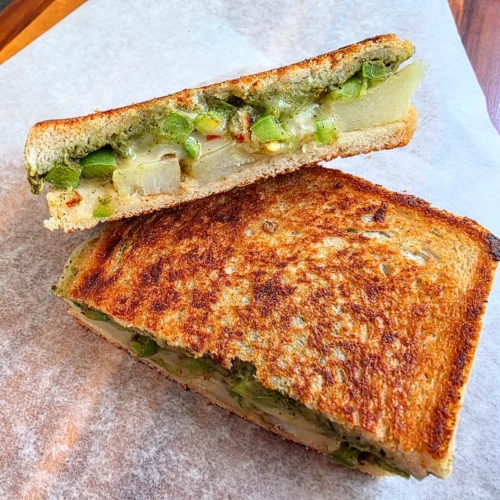 Grilled potato chutney sandwich cut in half, showing melted cheese, spiced potato slices and green chutney between toasted sourdough.